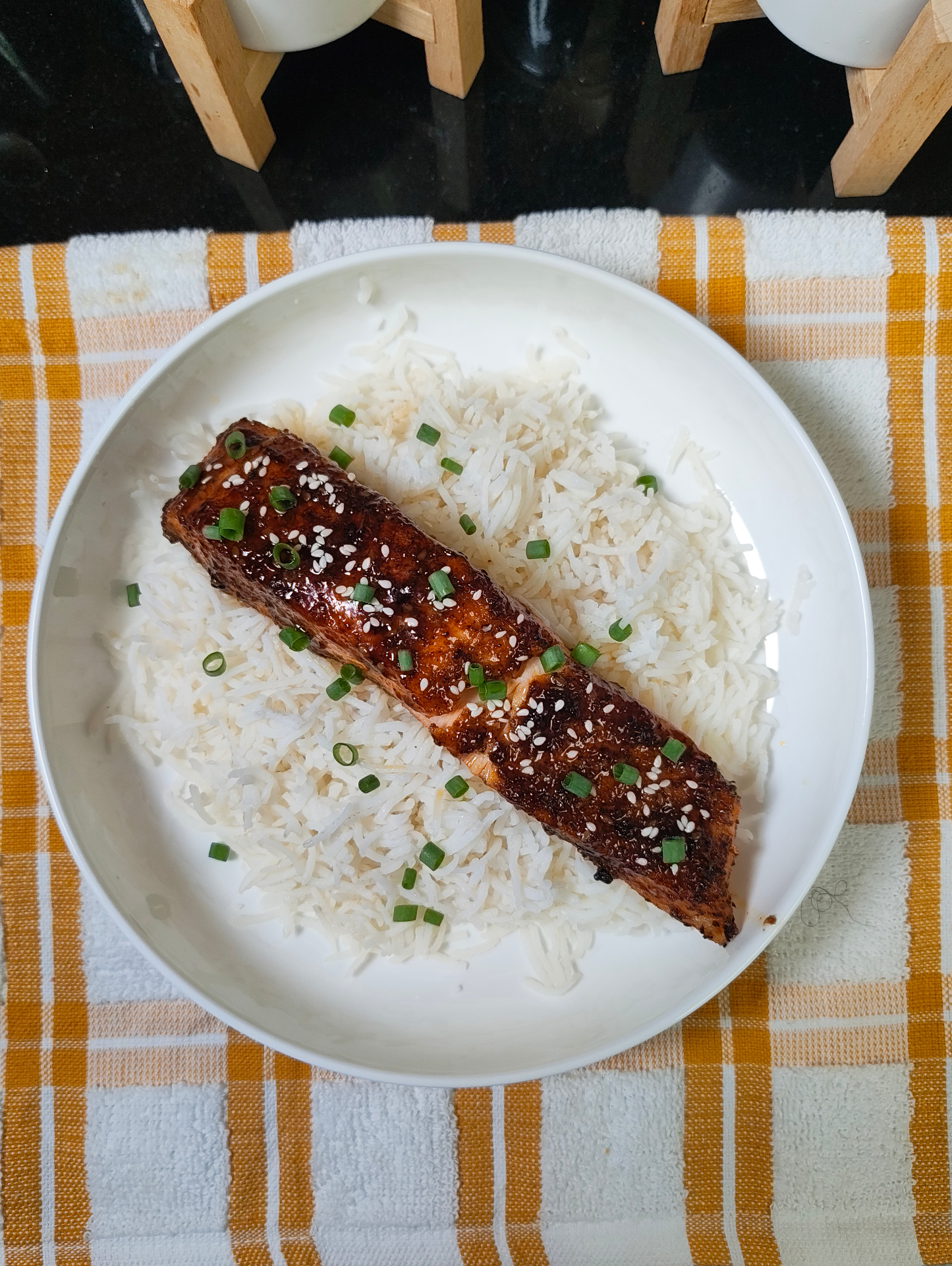 Overhead view of a glazed salmon fillet over white rice in a white bowl, sprinkled with sesame seeds and green onions, on a yellow checked cloth