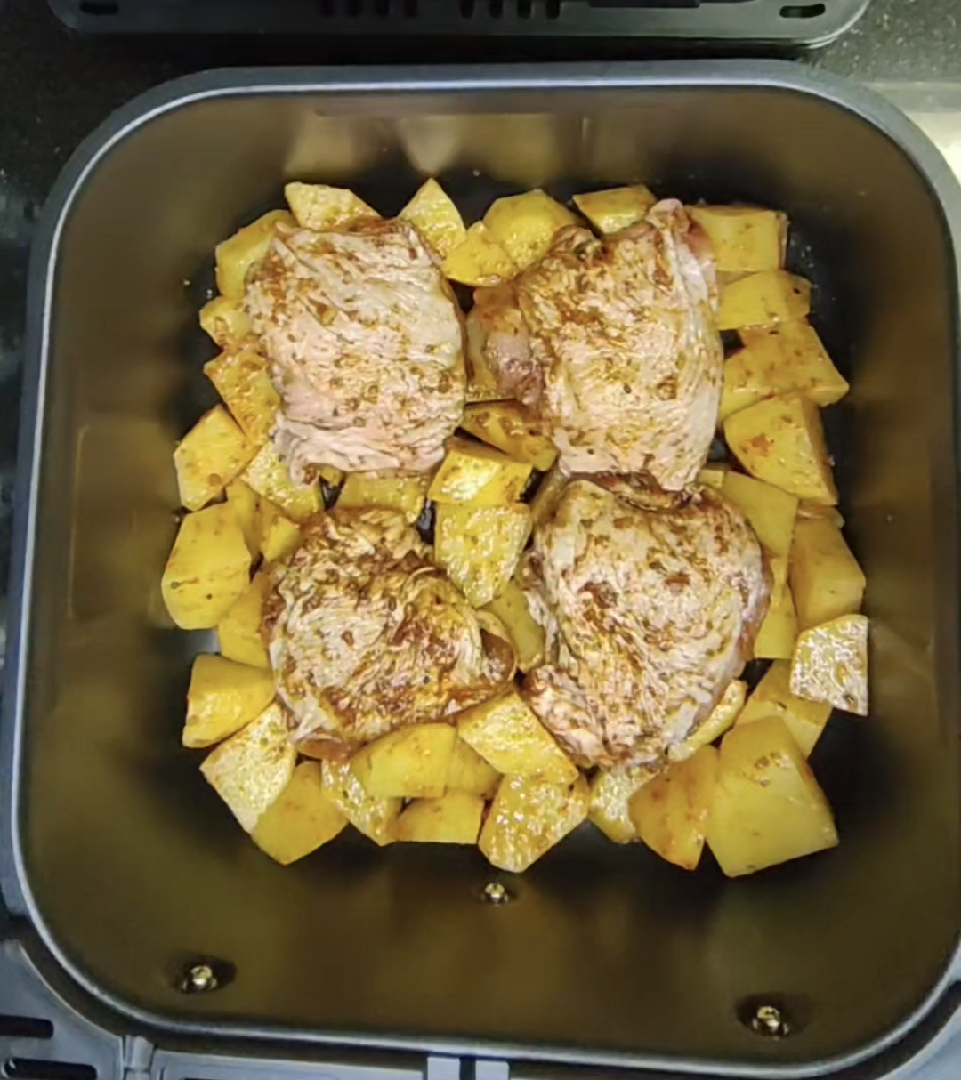 Four seasoned chicken thighs surrounded by potato chunks in an air fryer basket before cooking