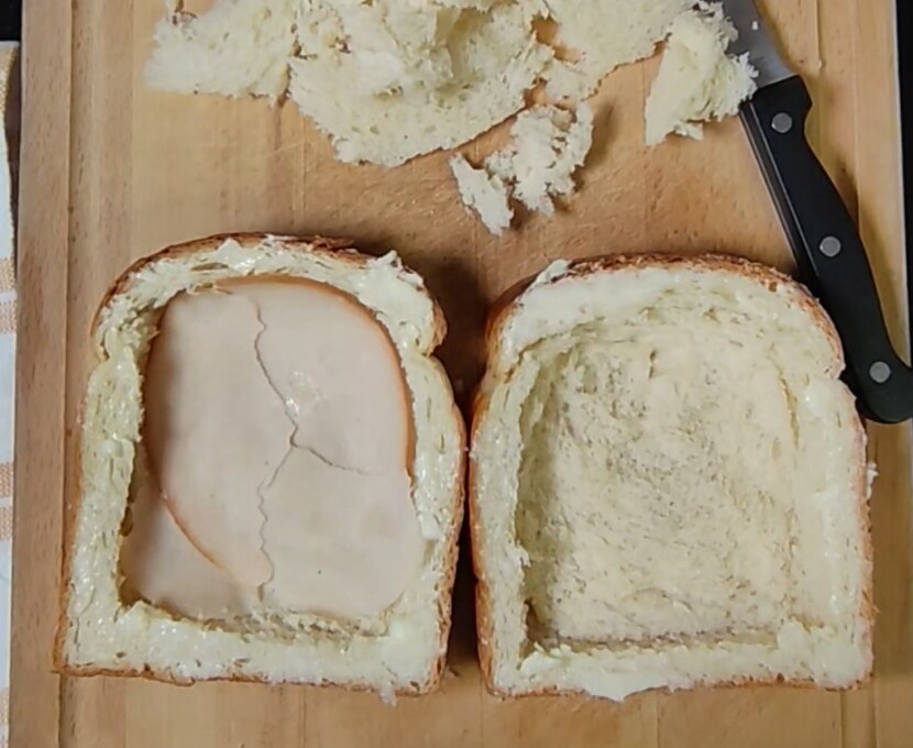 Two hollowed brioche slices on a wooden board, one lined with slices of turkey breast, with a knife and removed bread nearby