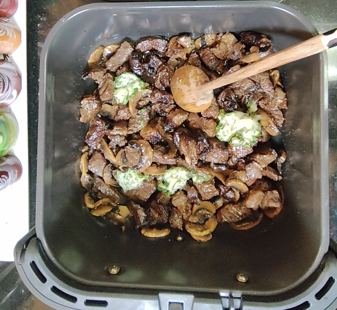 Cooked beef and mushrooms in the air fryer basket with dollops of parsley garlic butter and a wooden spoon