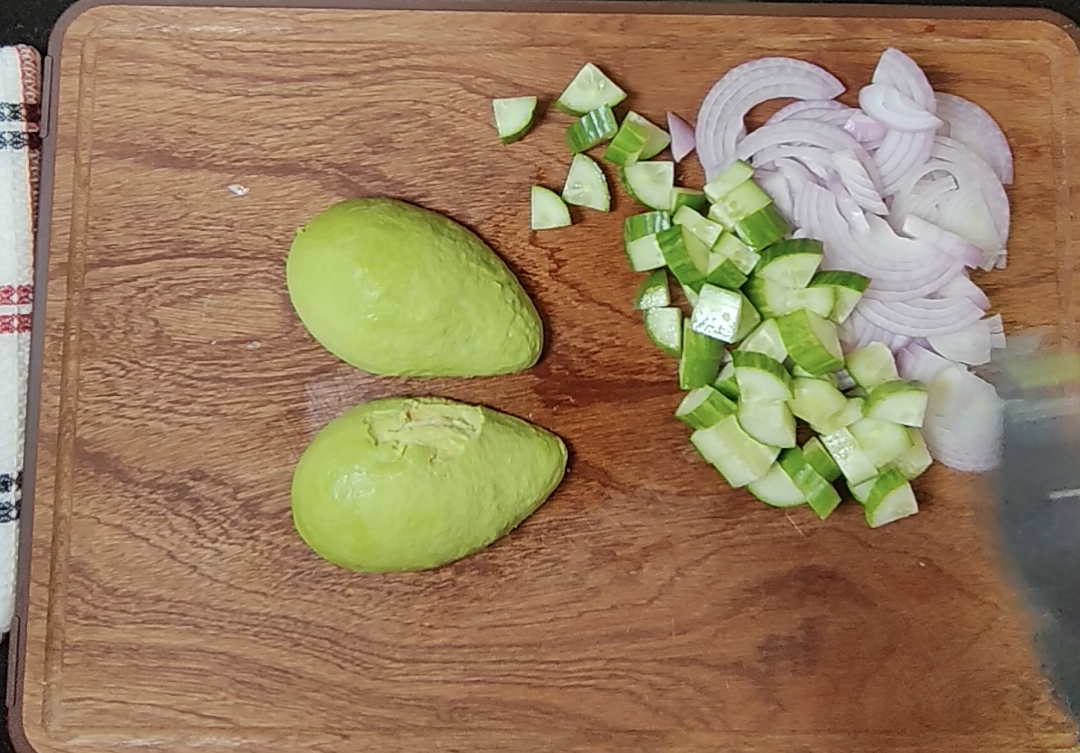 Halved avocado, chopped cucumber and sliced onion on a wooden chopping board