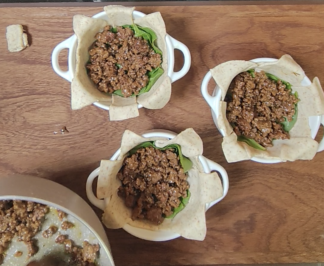 Three wholemeal pastry lined ramekins with fresh spinach and a first layer of cooked minced beef on a wooden board