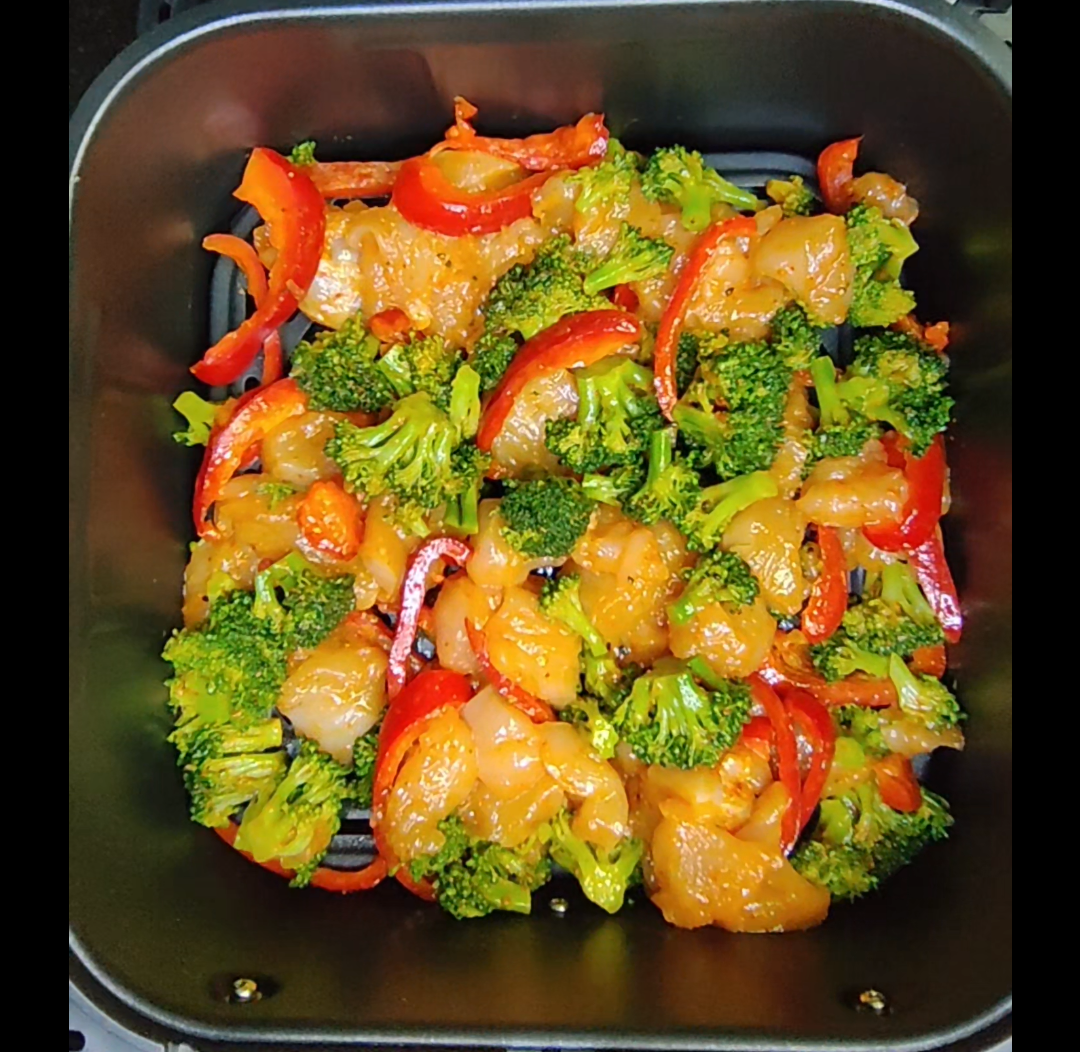 Uncooked chicken pieces, broccoli florets and red pepper spread across an air fryer basket before cooking