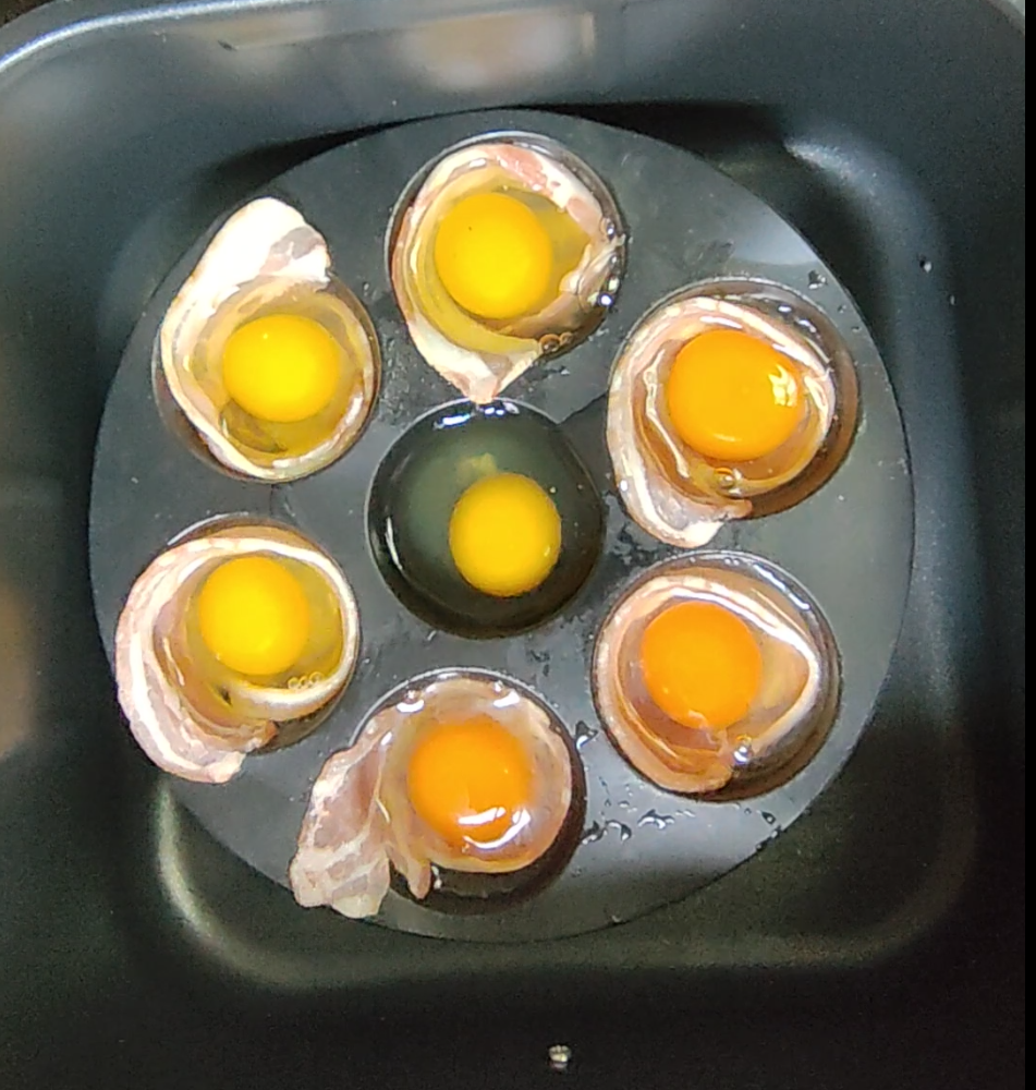 Raw eggs cracked into a bacon-lined silicone mould inside an air fryer basket