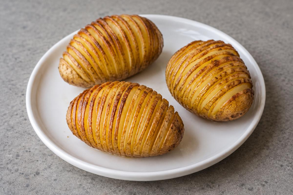 Editorial illustration of three golden Hasselback potatoes with closely spaced slices on a white plate