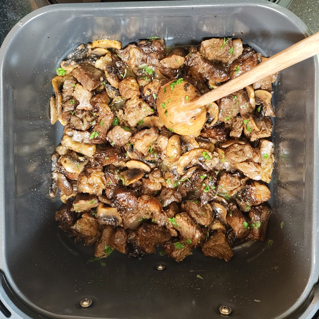 Browned beef and mushroom pieces with a wooden spoon in an air fryer basket