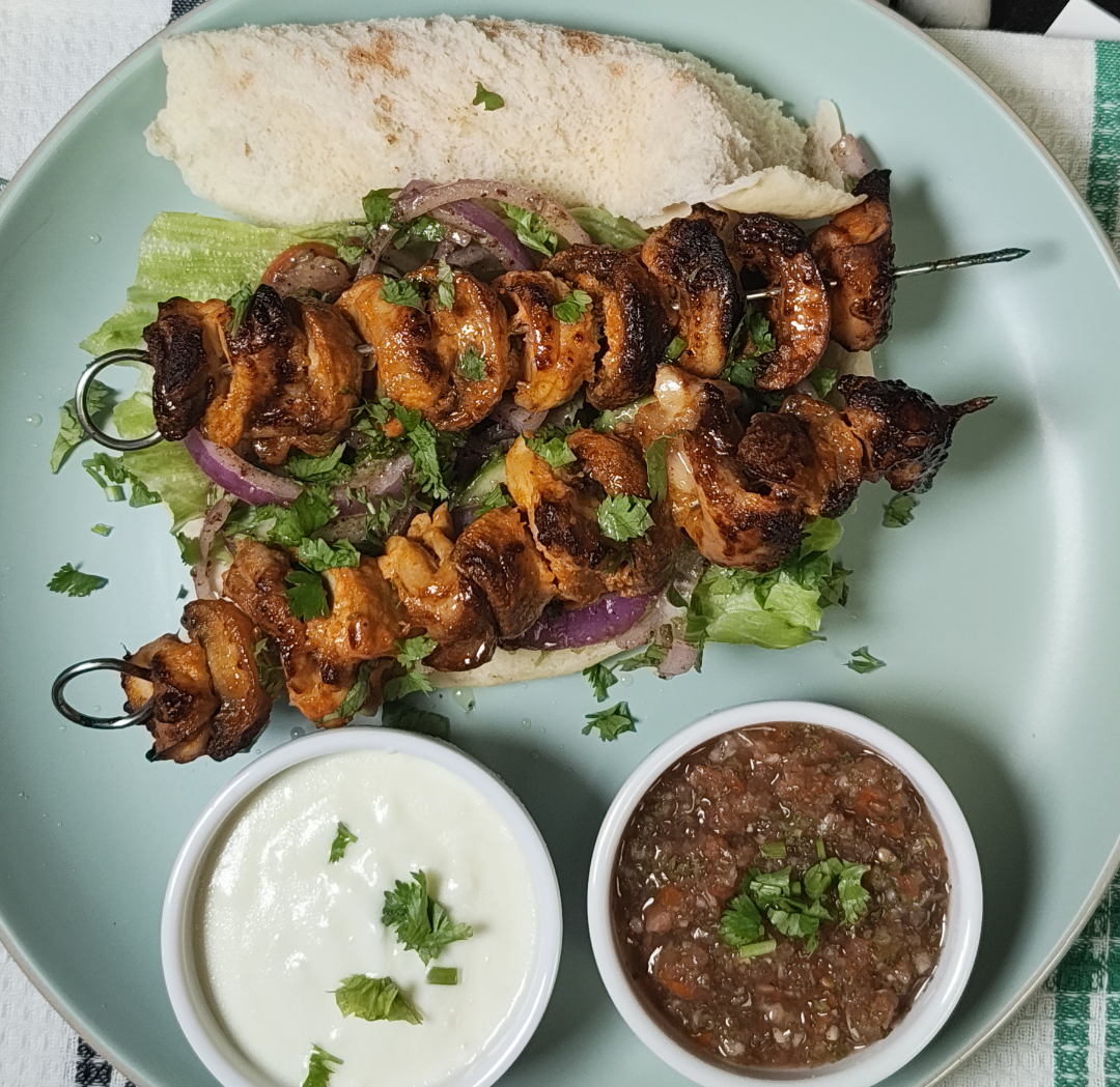 Cooked chicken tikka and mushroom skewers garnished with coriander, served with flatbread and two sauce bowls on a pale blue plate